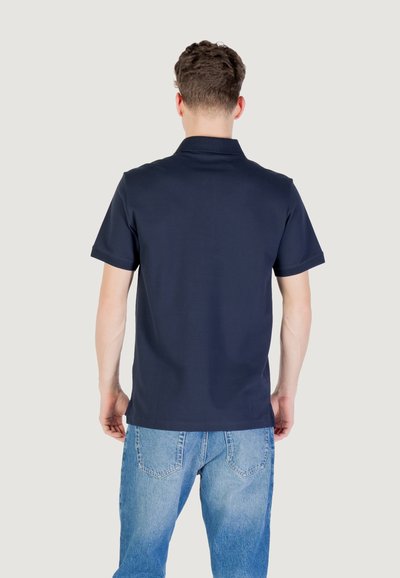 Temno modra polo majica z oblikovanim ovratnikom in kratkimi rokavi, v kombinaciji s svetlo modrimi kavbojkami. Gladka tkanina z minimalno teksturo.