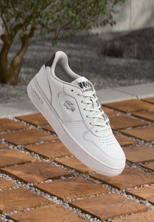 Adidași albi din piele cu o suprafață texturată, cutie perforată pentru degete, design curbat și un accent negru pe călcâi. Prezintă branding Lacoste.