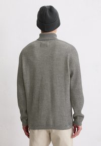 Uomo con un maglione a collo alto grigio a coste, pantaloni beige e berretto di maglia grigio scuro, di spalle contro un muro bianco semplice.