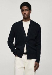 Námorníkový cardigan vyrobený z měkkého pleteného materiálu, s hlubokým V-výstřihem, pěti knoflíky na přední straně a žebrovanými manžetami. Nositý přes bílé tričko.