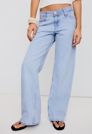 Person wearing light blue wide-leg jeans, cropped white top, black flip-flops, and chunky beige bracelets on left wrist.