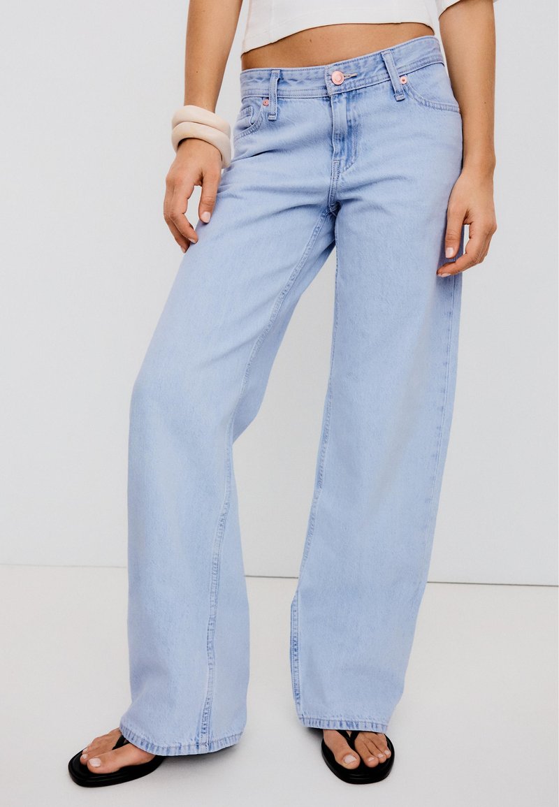 Personne portant un jean bleu clair à jambes larges, un top blanc court, des tongs noires et des bracelets beige épais au poignet gauche.