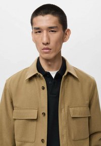 Tan button-up jacket with a collared neck, two chest pockets, and a textured fabric, layered over a black polo shirt.
