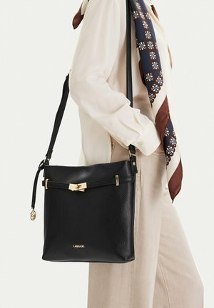 Mujer vestida con blusa color crema y pantalones beige, llevando un bolso de hombro de cuero negro Lasocki con cierre dorado y una bufanda estampada sobre el hombro.