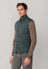 Gesteppte Weste in tiefem Teal mit Stehkragen und Knopfverschluss vorne. Trägt man über einem hellbraunen Rollkragenpullover mit strukturierter Oberfläche.