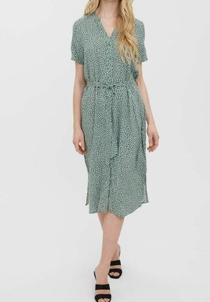 Nainen vihreässä midi-mekossa, jossa on valkoisia pilkkuja, lyhyet hihat, solmittava vyö ja mustat avoimet sandaalit yksivärisellä taustalla.