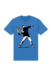 Osoba v černé mikině a bílých kalhotách připravena hodit kytici červených a zelených květin, vytištěnou na modrém tričku.