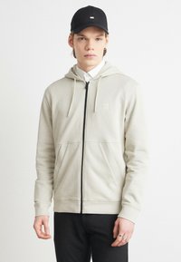 Felpa zip grigia chiara con cappuccio regolabile, tasche frontali e zip nera. Indossata con una maglietta bianca e un cappello nero.