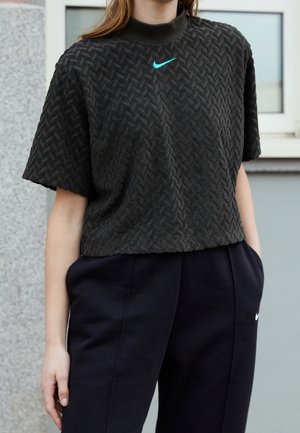 Person iført en tekstureret sort kortærmet top med turkis Nike-logo og sorte Nike-bukser, stående udendørs tæt på et vindue.