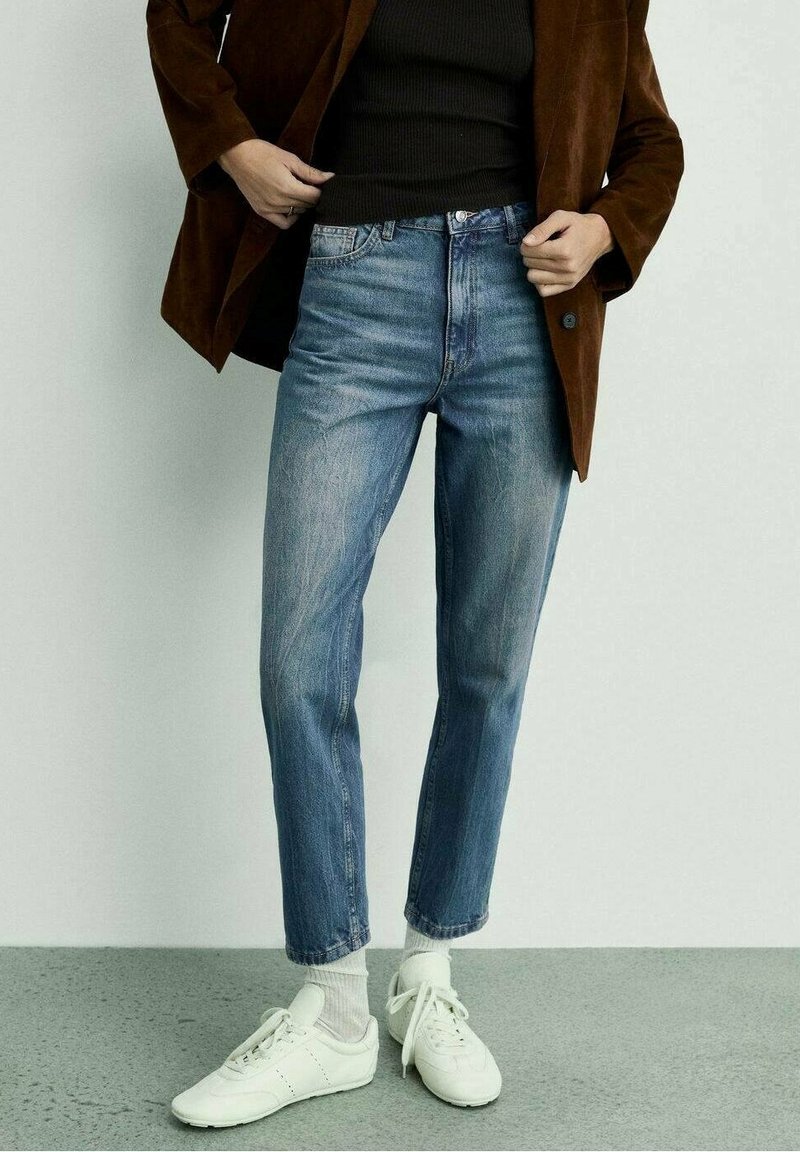 Jeans in denim azzurro chiaro con una vestibilità dritta, caratterizzati da leggere sfumature. Abbinati a un top scuro e scarpe da ginnastica bianche. Blazer marrone appoggiato sopra.