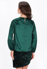 Grüne Satinbluse mit langen Ärmeln und einer Aussparung am Rücken, kombiniert mit einem dunkelgrünen Rock mit Pailletten, der eine strukturierte Oberfläche zeigt.