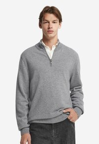 Grå zip-up tröja i mjukt stickat material, med ståkrage och ribbade mudd, tillsammans med en vit skjorta under.