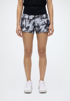 Modèle portant un short de running noir et gris à motif tie and dye, un haut noir et des baskets blanches sur un fond uni clair.