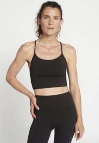 Donna con capelli ricci, in un top corto nero e leggings a vita alta, in piedi con una mano sulla coscia contro uno sfondo bianco.