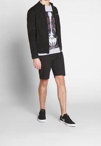 Veste en denim noir avec fermeture éclair à l'avant, finitions côtelées sur les manches, t-shirt graphique gris, shorts noirs et baskets noires avec semelles blanches.
