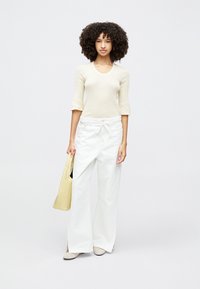 Top en maille côtelée beige clair à manches trois-quarts, associé à un pantalon large blanc. Le modèle tient un sac fourre-tout jaune pâle.