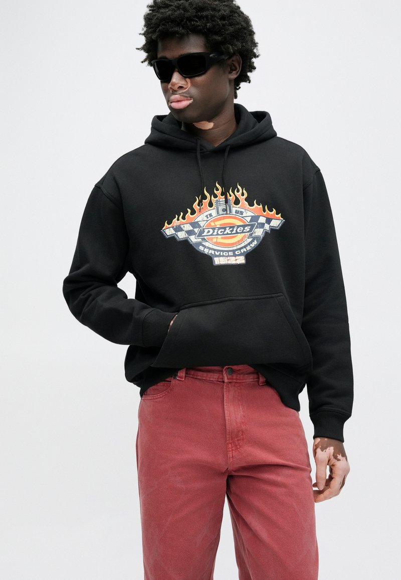 Black hoodie with a graphic featuring flames and the word "Dickies," paired with red pants. Model wearing sunglasses, hands in pockets.