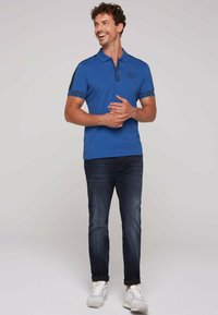 Camiseta polo azul con cuello con cremallera, detalles en negro en los hombros y logo en la manga. Combinada con vaqueros oscuros y zapatillas blancas.