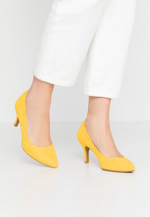Escarpins jaunes en daim à talon haut, avec un bout pointu et un talon fin, associés à un pantalon blanc cropped. Texture lisse, design minimaliste.
