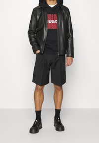 Černá kožená bunda přes černou mikinu s červeným logem. Volné černé kraťasy a černé platformové sneakers dotvářejí celkový vzhled.