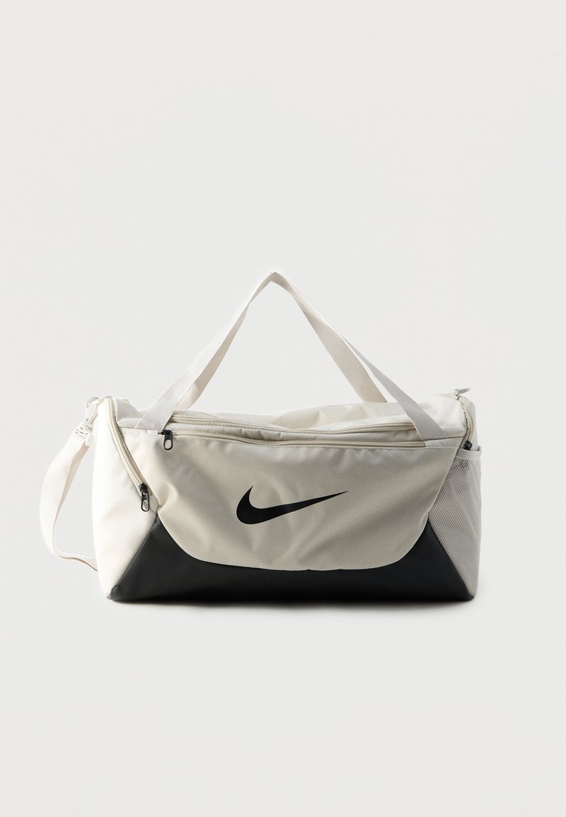 Λευκή και μαύρη τσάντα duffel Nike με διπλές λαβές, φερμουάρ σε διαμερίσματα και πλαϊνή θήκη από δίχτυ σε μονόχρωμο φόντο.