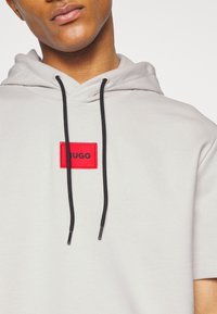 Sudadera con capucha de color gris claro con un parche rectangular rojo con el logo "HUGO", que presenta cordones negros y un material de algodón suave y texturizado.