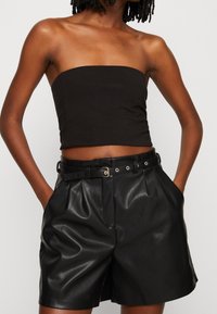 Crop top noir sans manches associé à un short en cuir noir taille haute. Le short présente des plis et une ceinture avec des éléments en métal.