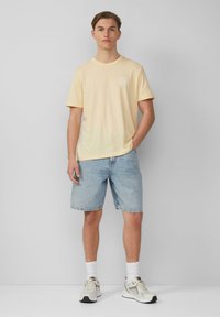 T-shirt en coton jaune avec un col rond, coupe décontractée, associée à un short en denim bleu clair et des chaussettes blanches avec des baskets grises.