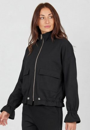 Žena s dlhými hnedými vlasmi, ktorá nosí čiernu bundu na zapínanie so širokými prednými vreckami a elastickými volánikovými manžetami, pozerá sa doľava.