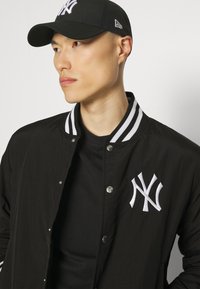Chaqueta bomber negra con cuello de rayas blancas, cuenta con botones a presión y un logo de NY bordado. Lleva una camiseta negra debajo y una gorra a juego.