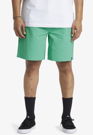 Pantalones cortos verdes de un tejido ligero y texturizado con una etiqueta negra de contraste. Combinados con calcetas negras y zapatillas negras.