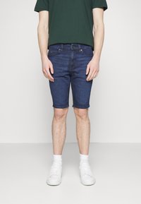 Osoba stojí v tmavě zeleném tričku a tmavých džínových šortkách, doplněná bílými teniskami a ponožkami, na jednobarevném pozadí.