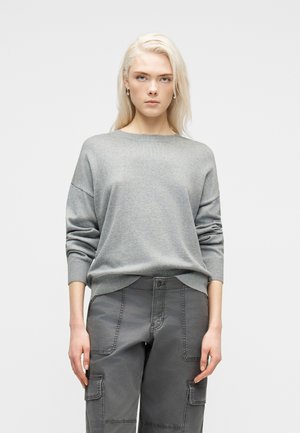 Kobieta o długich blond włosach, ubrana w szary sweter i szare spodnie typu cargo, stojąca na tle jednolitego jasnego tła.