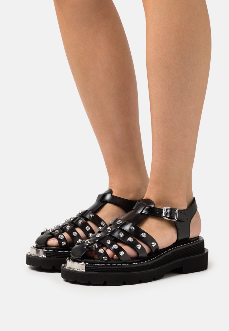Sandales en cuir noir avec plusieurs brides ornées de clous argentés, semelles épaisses et bulkys, et brides à boucle à la cheville, portées aux pieds sur fond blanc.