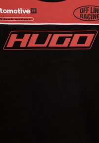 T-shirt in cotone nero con testo rosso e bianco, inclusa la scritta "HUGO" in evidenza. Include dettagli grafici e loghi di accento.