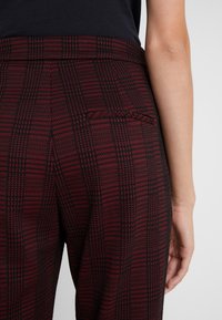 Vue arrière d'une personne portant un pantalon à carreaux rouge foncé et noir avec une ceinture ajustée et une petite poche passepoilée à l'arrière.