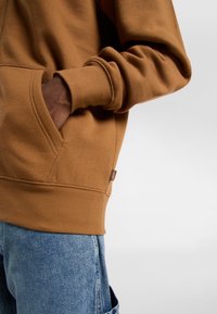 Brown zip-up hoodie with ribbed cuffs and hem, featuring a front pocket and soft texture, paired with denim jeans.