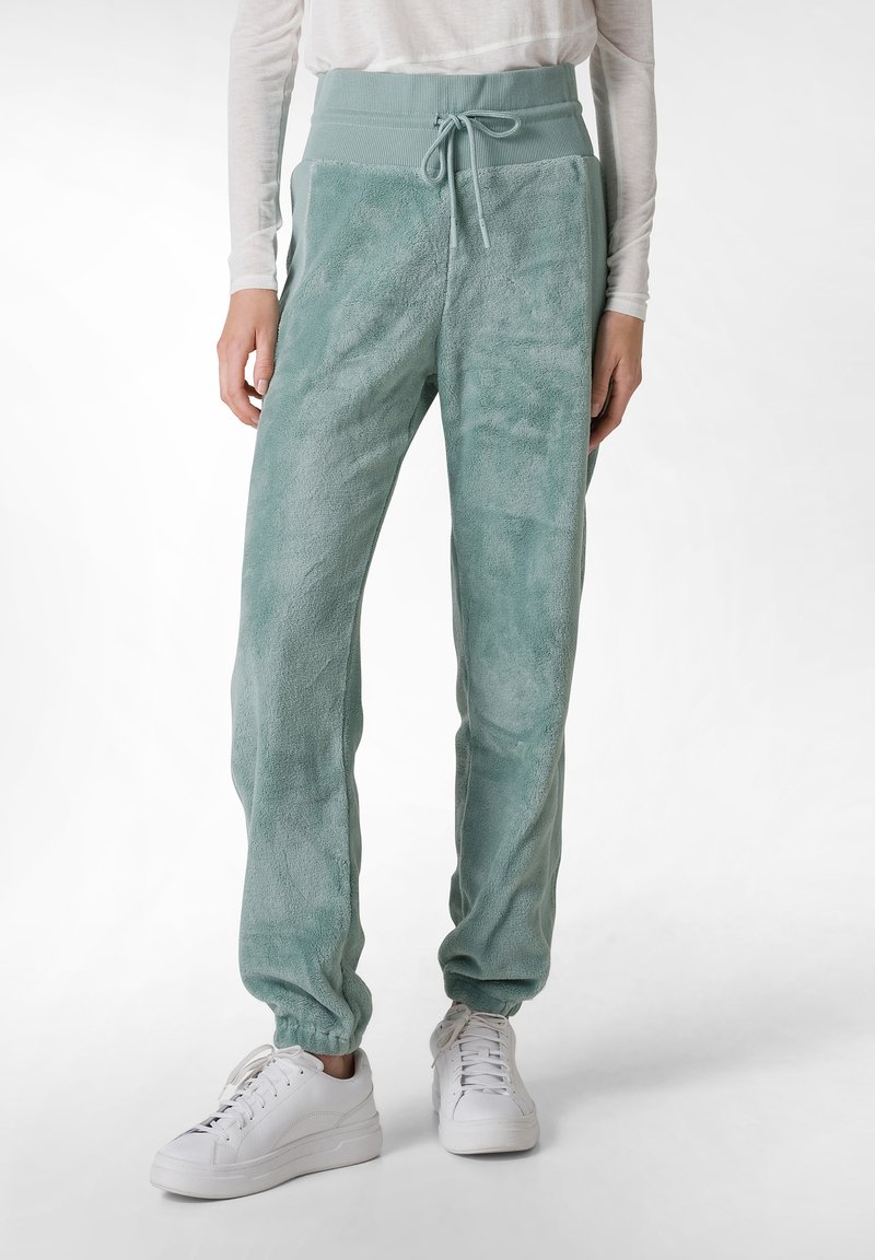 Fleece joggers i dæmpet grøn med snøre i taljen, elastiske manchetter og en blød tekstur, parret med hvide sneakers.