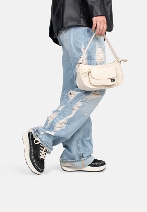 Bolso de piel blanco con un bolsillo frontal, sostenido por una persona que lleva unos jeans claros desgastados y zapatillas negras con detalles en blanco.