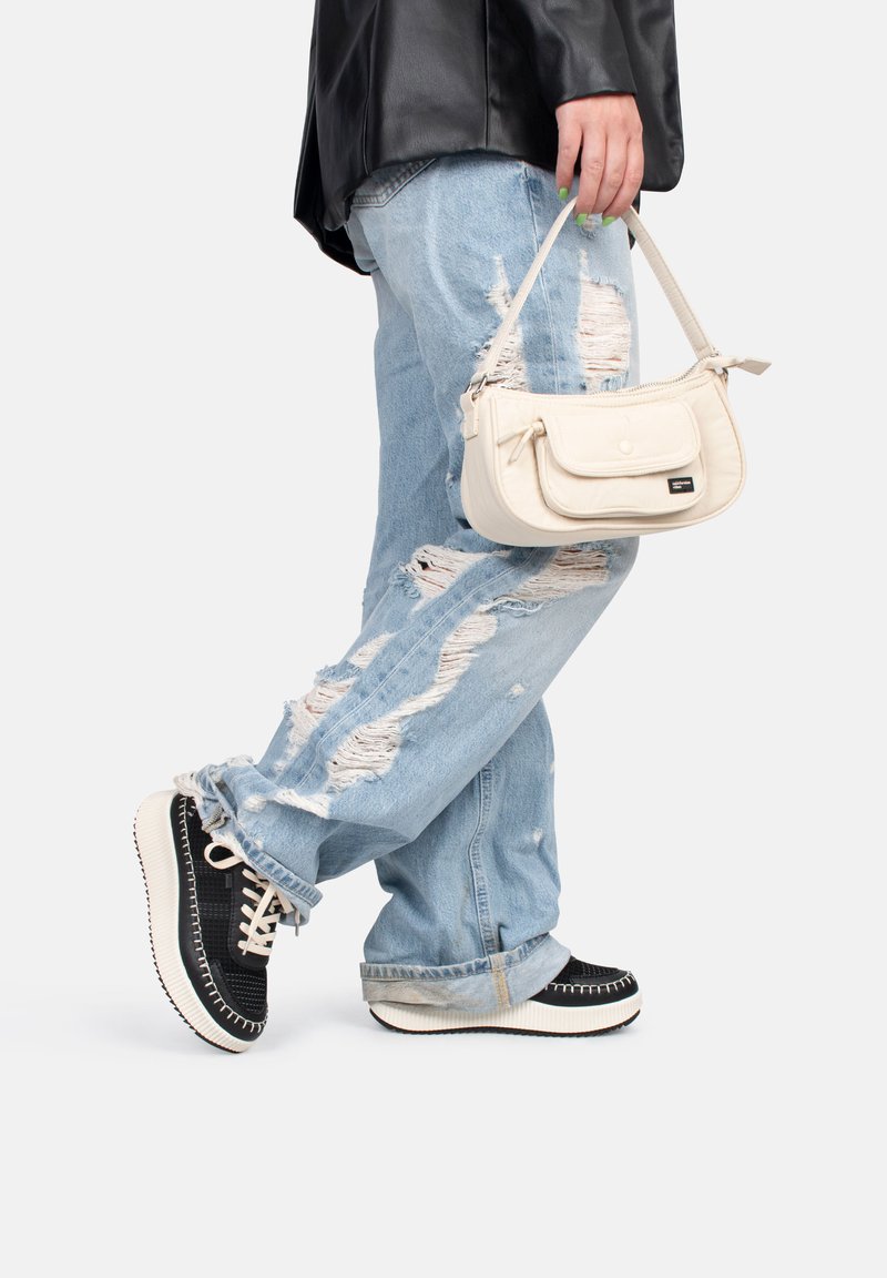 Bolso de piel blanco con un bolsillo frontal, sostenido por una persona que lleva unos jeans claros desgastados y zapatillas negras con detalles en blanco.