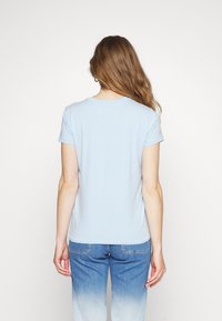 Světle modré tričko s krátkým rukávem vyrobené z měkké látky, s kulatým výstřihem a přiléhavým střihem. Zadní strana má hladkou texturu.