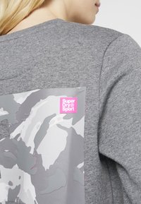 Felpa grigia con un design mimetico testurizzato in diverse sfumature di grigio, con un brillante logo "Super Dry Sport" in rosa nell'angolo.