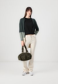 Cardigan verde y gris sobre una blusa negra, combinado con jeans cremosos y zapatillas blancas. Sosteniendo un bolso de pana verde oliva con bolsillos.