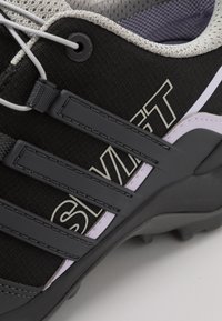 Zapato deportivo negro con parte superior de tela texturizada, tiras grises, forro de malla y suela de goma con tacos para mayor tracción.