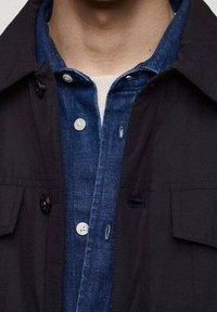 Czarna zewnętrzna kurtka z zapinanym na guziki kołnierzem, noszona na niebieskiej jeansowej koszuli z dwoma guzikami i teksturowaną powierzchnią.