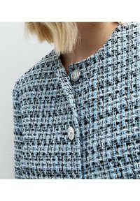 Tweedjacka i ljusblått med en strukturerad mönstring. Har silverknappar och en rund halsringning. Nära vy av tygdetalj.