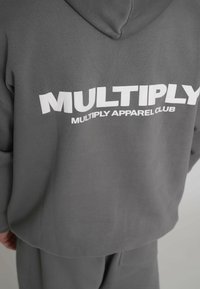 Sudadera gris con capucha que presenta texto blanco en negrita "MULTIPLY" y "MULTIPLY APPAREL CLUB" en la parte trasera, diseño casual con textura suave.