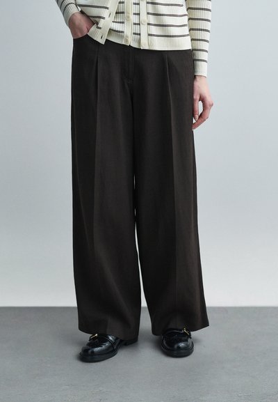 Personne portant un pantalon large ample marron foncé, des mocassins en cuir noir et un cardigan blanc à rayures noires, debout sur un sol gris.