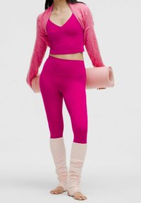 Jasnofuksjowy strój sportowy z krótkim topem i legginsami o wysokim pasie, połączony z jasnoróżowymi ocieplaczami na nogi i matą do jogi.
