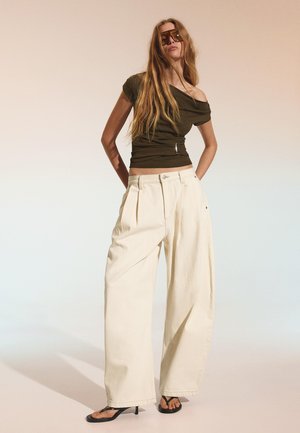 Femeie cu păr lung, purtând ochelari de soare pătrați supradimensionați, bluză olive pe umeri, pantaloni largi crem și sandale negre cu toc, stând în fața unui fundal bej.